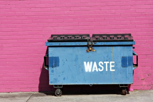 A large blue industrial waste bin with a hinged lid and metal handle, positioned on a sidewalk in front of a vibrant pink brick wall. The bin has visible dirt marks and wear, with the word 'WASTE' painted in white on its side. It stands on four small wheels, with some wheels and metal supports visible beneath. The setting suggests an urban environment, possibly near commercial premises in Hammersmith, with a concrete pavement extending from the base of the wall. The bright pink background contrasts sharply with the utilitarian appearance of the bin, highlighting its practical role in waste collection or rubbish removal services provided by Commercial Waste Removal Hammersmith. The scene is well-lit, indicating daylight, and the overall composition emphasizes the bin as a functional element amidst a colourful, urban landscape.