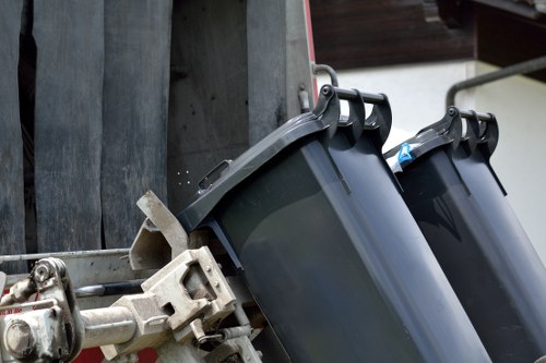 The image shows two dark grey wheelie bins with black lids, positioned side by side and slightly angled, attached to a metal waste container on a residential driveway or pavement. The bins are made of durable plastic with textured surfaces, and their lids are closed. The waste container to which they are attached has a metallic, slightly weathered surface, and is secured with a metal bracket or latch system. In the background, there are vertical wooden panels and a section of a building structure, indicating a typical urban or suburban setting. The lighting appears natural, likely daylight, highlighting the details of the plastic bins and metal container. This scene relates to rubbish removal services, such as those provided by Commercial Waste Removal Hammersmith, operating in the local area, including Hammersmith and surrounding postcodes.