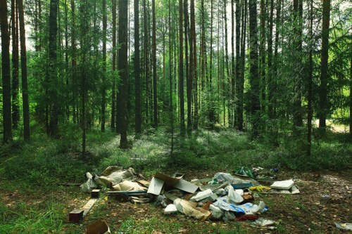 A natural woodland scene during daylight hours showing tall, slender trees with green leaves and straight trunks, creating a dense canopy overhead. The forest floor is covered with a mix of grass, small plants, and patches of soil. In the foreground, there is a pile of mixed waste debris, including cardboard boxes, plastic bags, and various packaging materials arranged haphazardly on the ground, indicating illegal dumping. The waste appears weathered and partially decomposed, contrasting with the lush, untouched environment behind it. This image highlights the issue of illegal waste disposal in natural areas within or near Hammersmith, suggesting the need for professional rubbish removal services like those provided by Commercial Waste Removal Hammersmith to manage such issues efficiently and maintain local environmental standards.