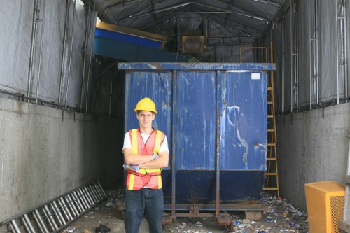 A young male waste management worker wearing a yellow safety helmet and a high-visibility vest with orange and yellow stripes stands with arms crossed in front of a large, blue industrial waste container inside a covered industrial area. The container has a weathered surface with visible scratches and dents, and is situated on a concrete surface scattered with small bits of debris. To the right of the container, there is a yellow ladder leaning against the side, and a yellow waste bin is visible at the bottom right of the image. The background shows the metal framework of the covered structure, with lighting that illuminates the scene uniformly, highlighting the textures of the container and the environment typical for waste collection and rubbish removal at a commercial or industrial site in London’s Hammersmith area. The image represents the services offered by Commercial Waste Removal Hammersmith, focusing on rubbish disposal and waste management.