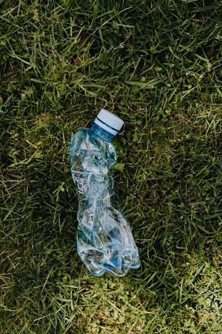A crumpled, transparent plastic water bottle with a white screw cap lies on a patch of green grass, surrounded by uneven blades of grass and small weed-like plants. The bottle appears partially flatte